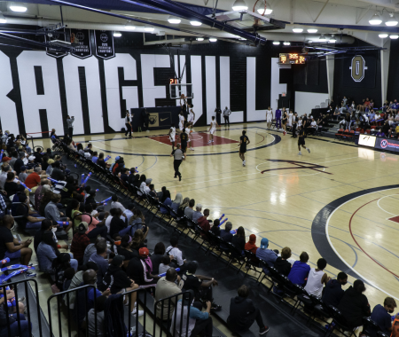 A backetball game is being played in front of a large crowd on an indoor court with Orangeville written on the wall in large white letters.